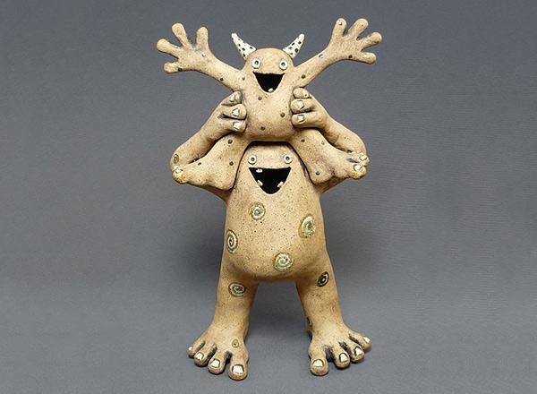 Two hand made ceramics creatures acting silly, one on the shoulders of the other.